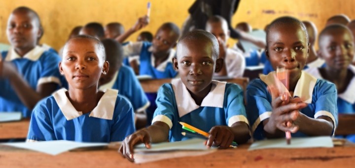 young girls in blue uniforms sit in a classroom holding pencils and papers.